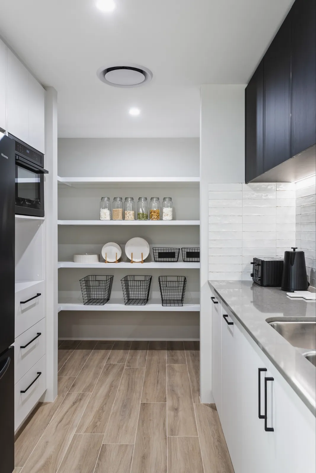 Butler's pantry with white open shelving holding glass jars, plates and wire baskets, beside a benchtop with a sink, toaster and kettle